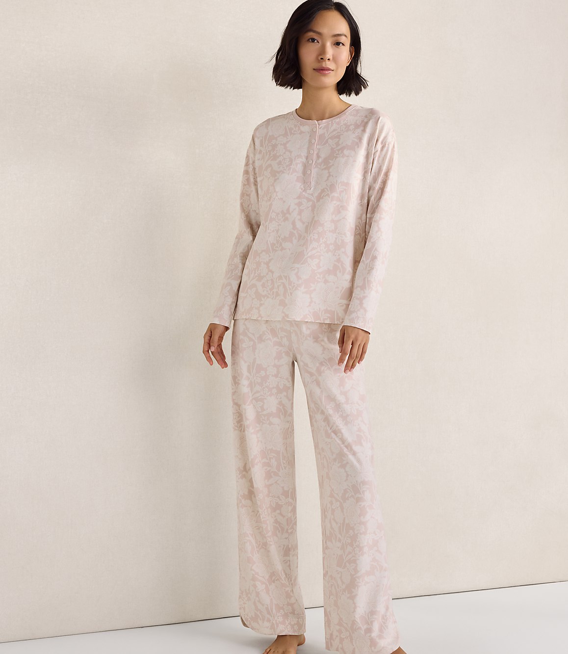 A woman wearing a light pink floral pajama set stands against a plain, neutral-toned wall.