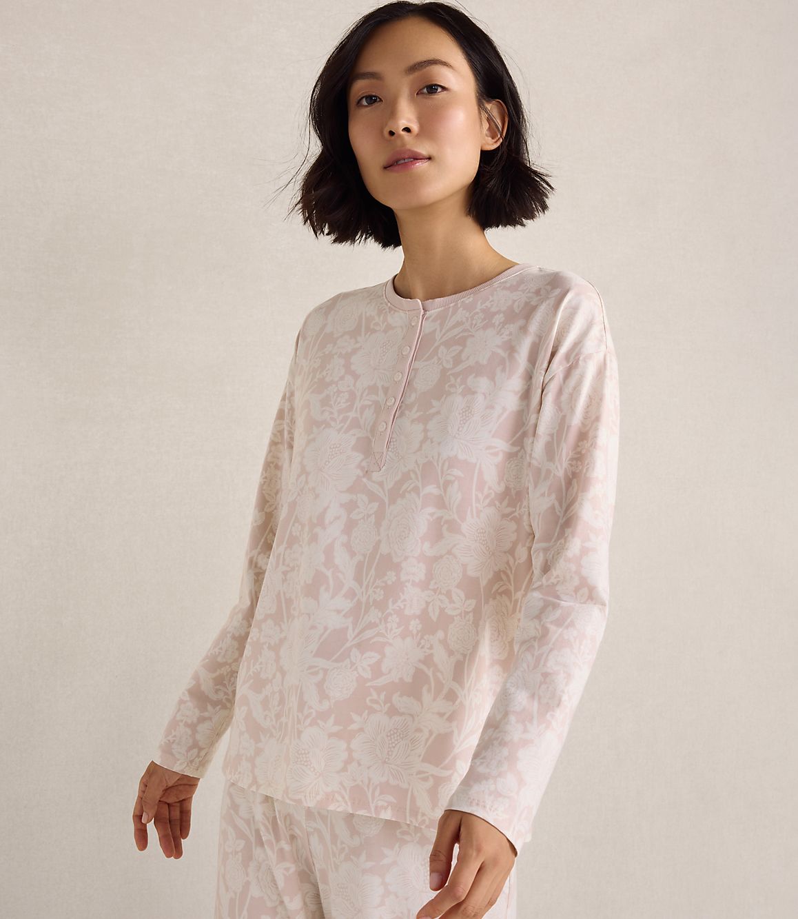 A woman wearing pink floral pajamas stands against a plain, neutral-toned background.