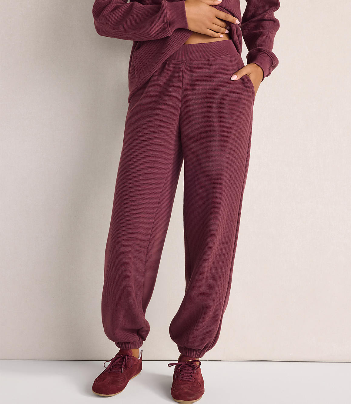 A person wearing a maroon textured lounge set and matching sneakers stands against a plain, light-colored wall.