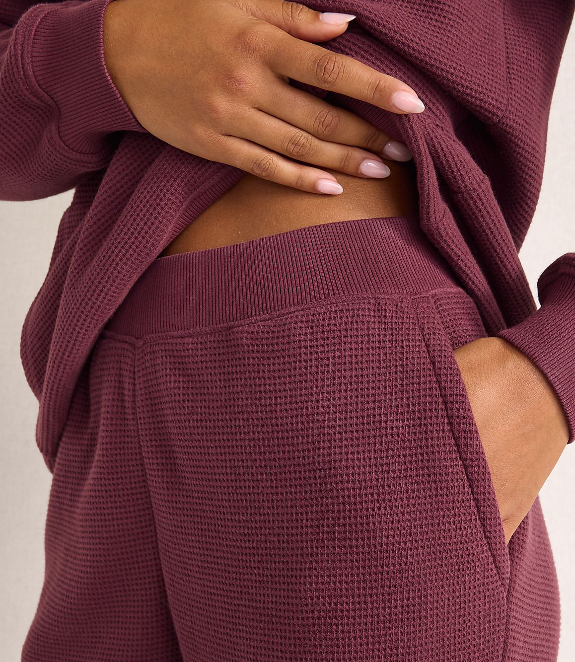 A person's hand rests on their stomach while wearing a maroon waffle-knit lounge set against a plain, light-colored background.
