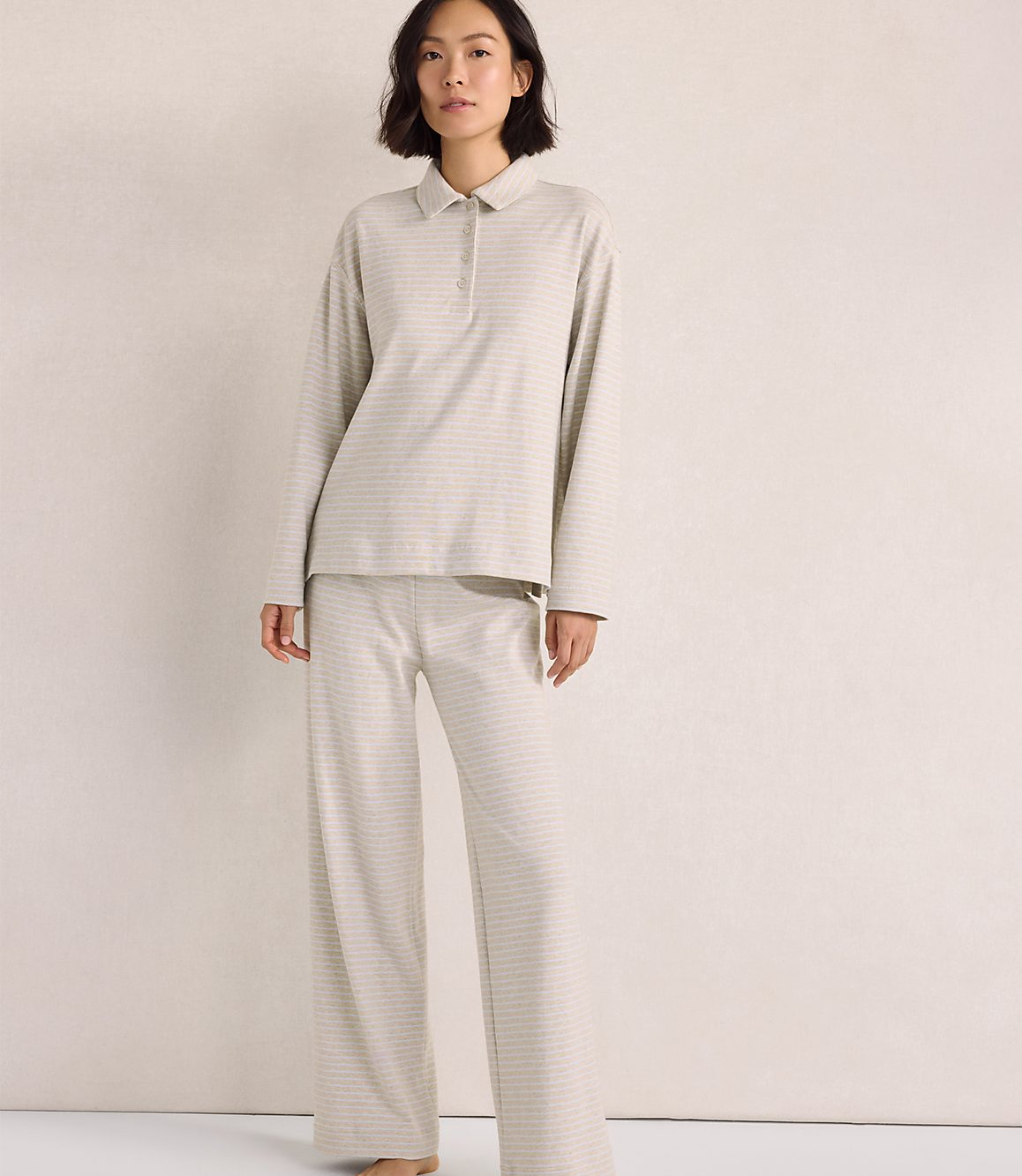 A woman wearing a light-colored, striped long-sleeved polo shirt and matching wide-leg pants stands against a plain, neutral-toned wall.
