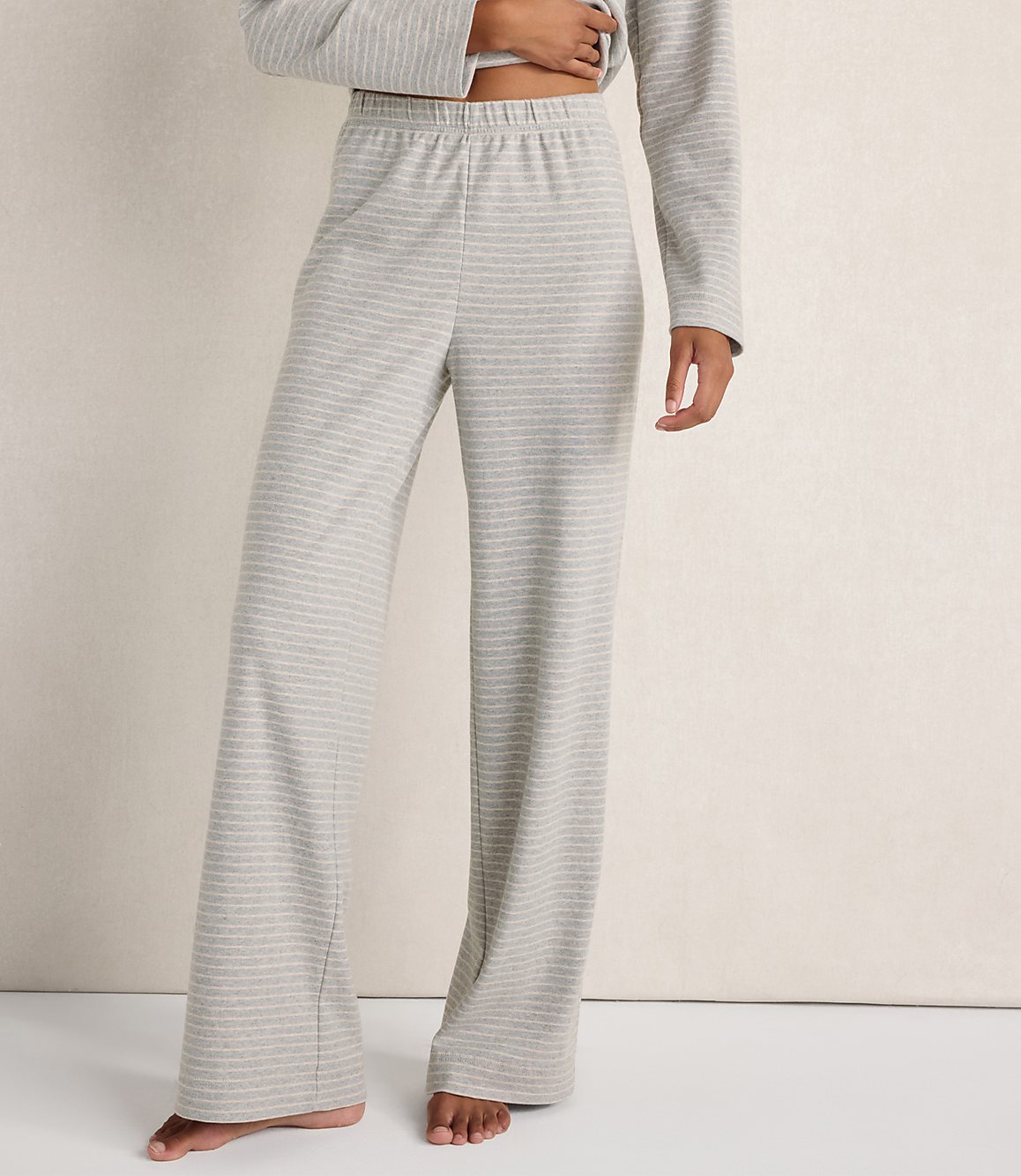 A person wearing grey striped lounge pants and a matching top stands against a plain, light-colored wall.