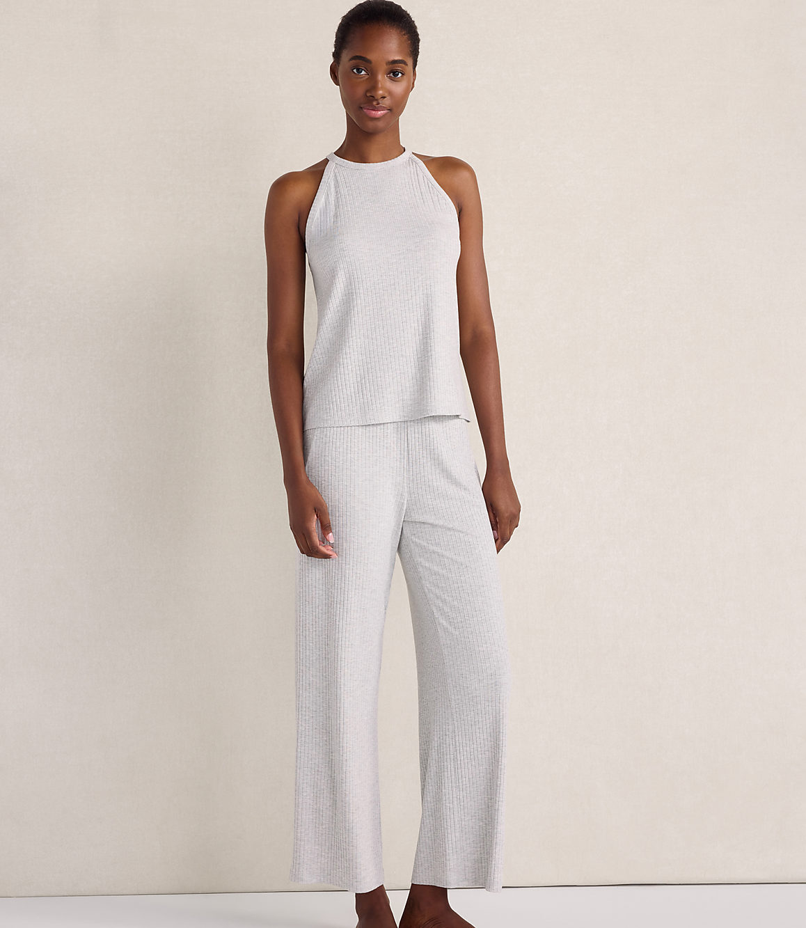 A woman wearing a light grey ribbed sleeveless top and matching wide-leg pants stands against a plain, neutral-toned wall.