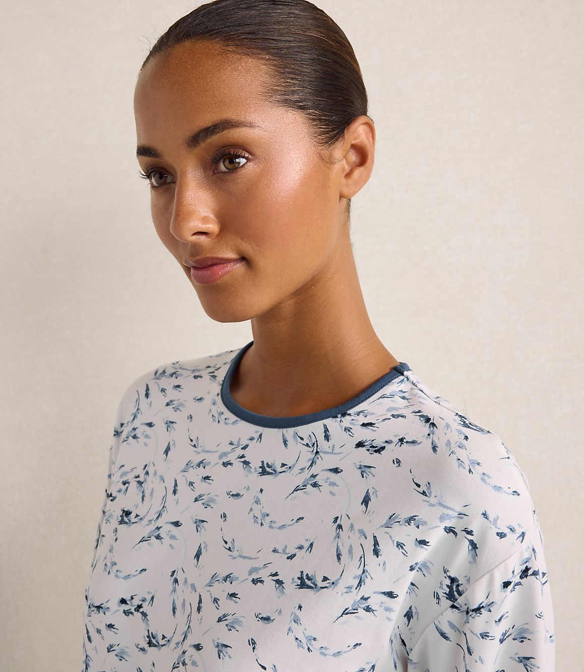 A woman with slicked-back hair wearing a white and blue patterned shirt stands against a plain, light-colored wall.