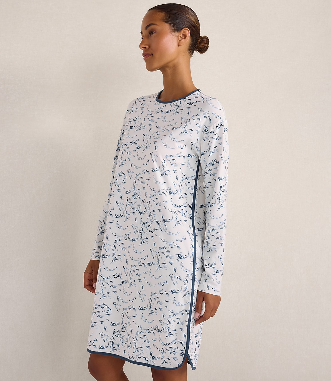 A woman wearing a white long-sleeved dress with a blue floral pattern stands against a plain, light-colored wall.