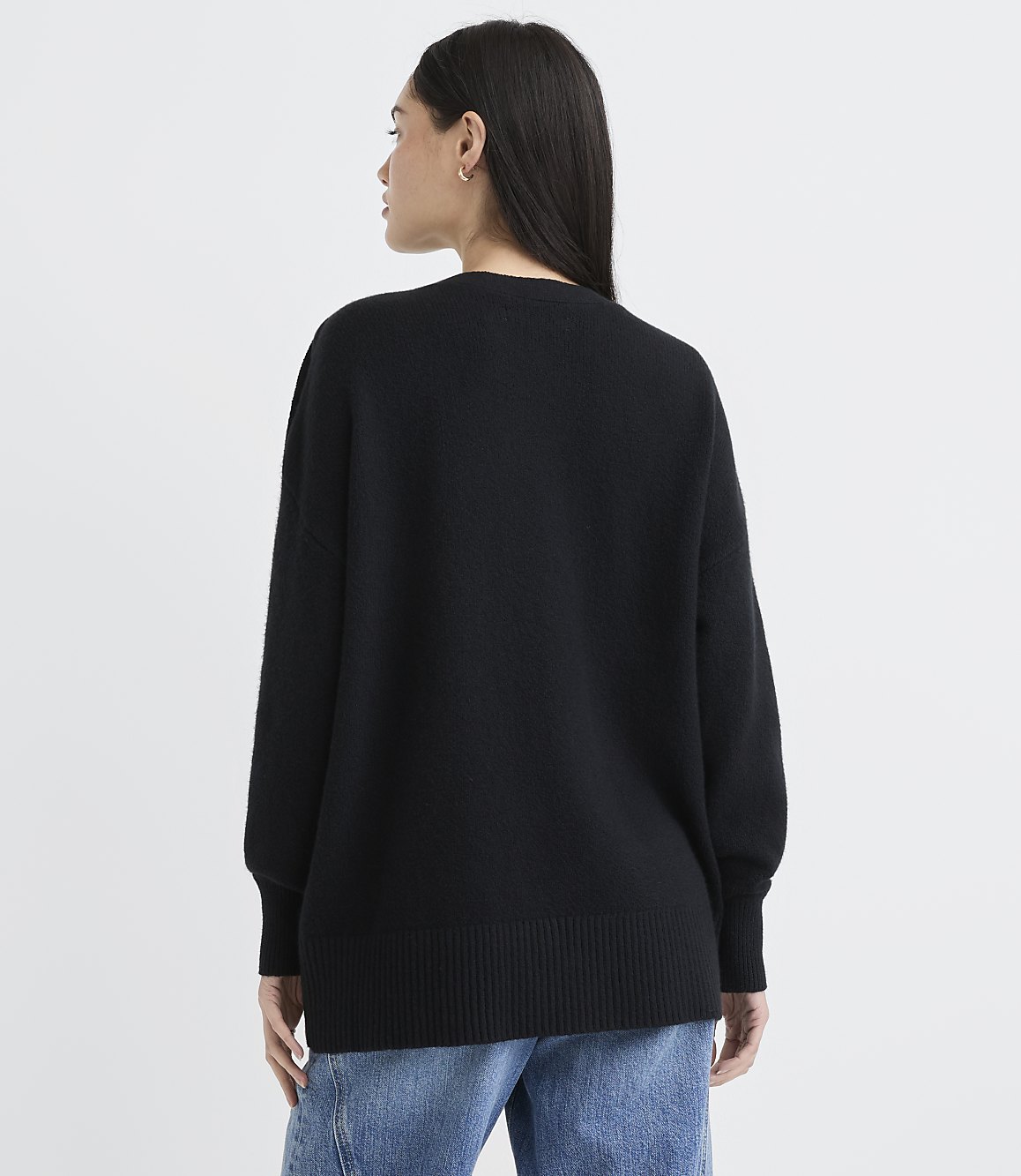 A woman wearing a black oversized sweater and blue jeans stands with her back to the camera against a plain white background.