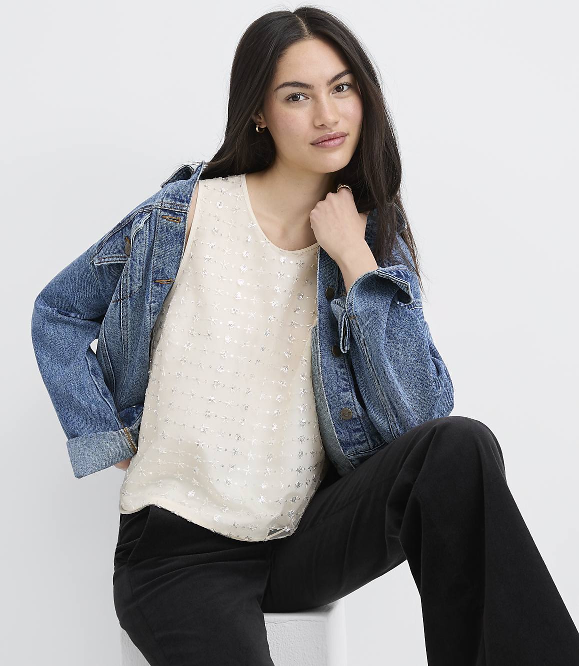 A woman wearing a white sequined top, a blue denim jacket, and black pants sits against a plain white background.