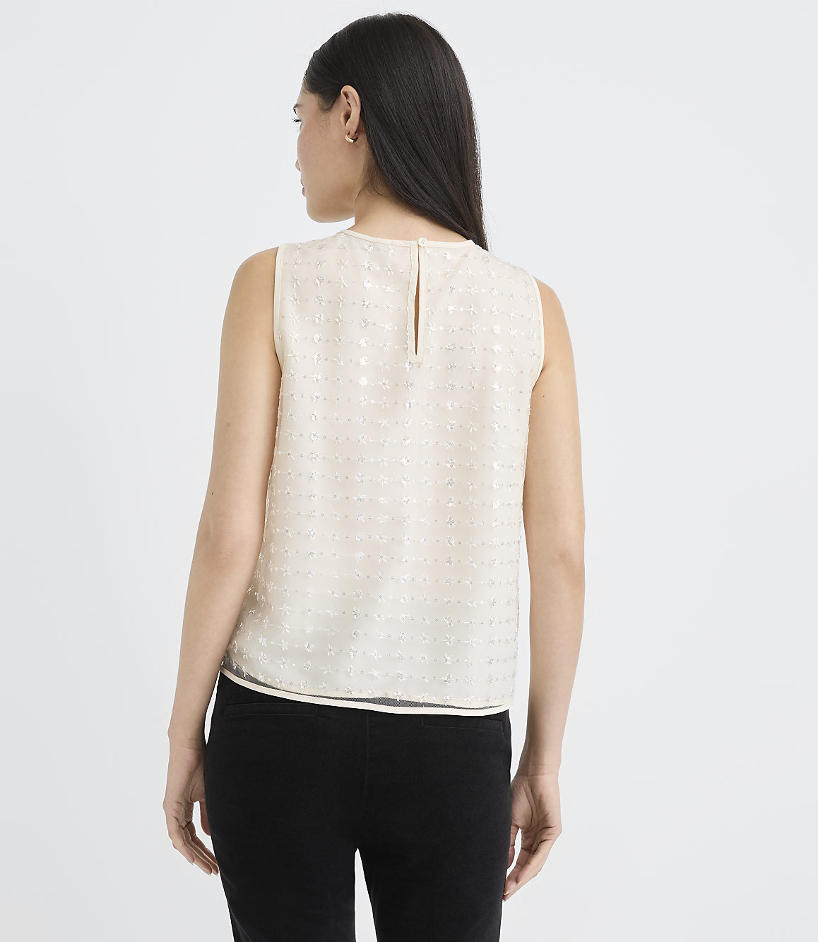 A woman wearing a sleeveless white textured top and black pants stands against a plain white background.