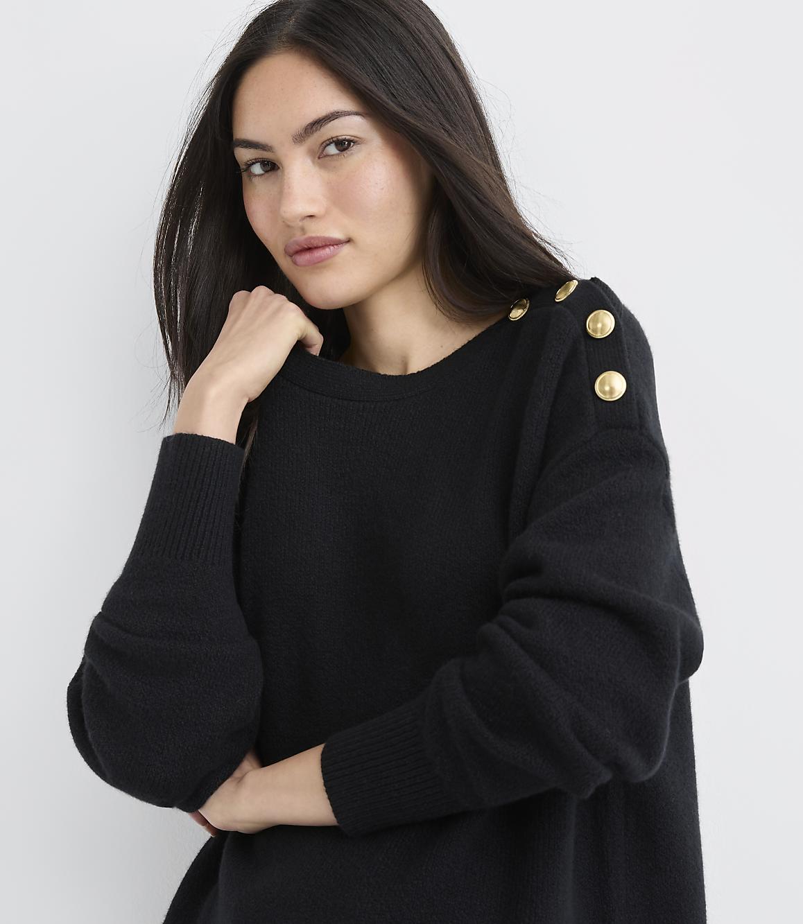 A woman wearing a black sweater with gold buttons on the shoulder poses against a plain white background.