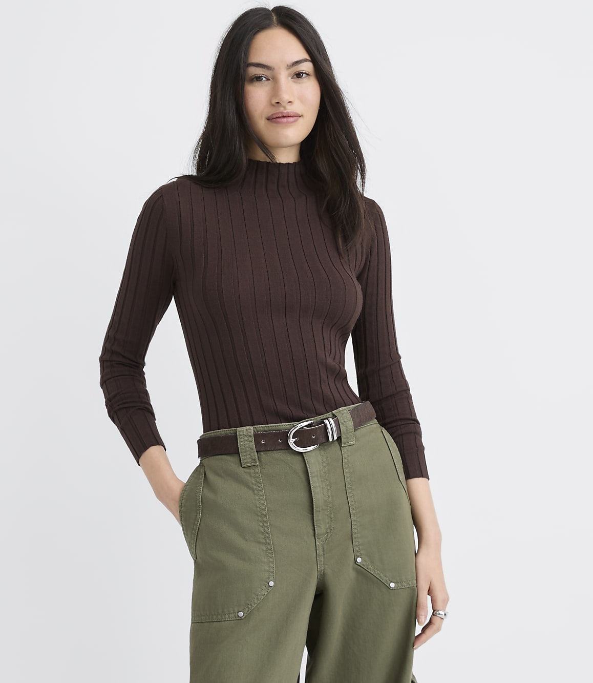A woman wearing a brown ribbed turtleneck and olive green pants stands against a plain white background.