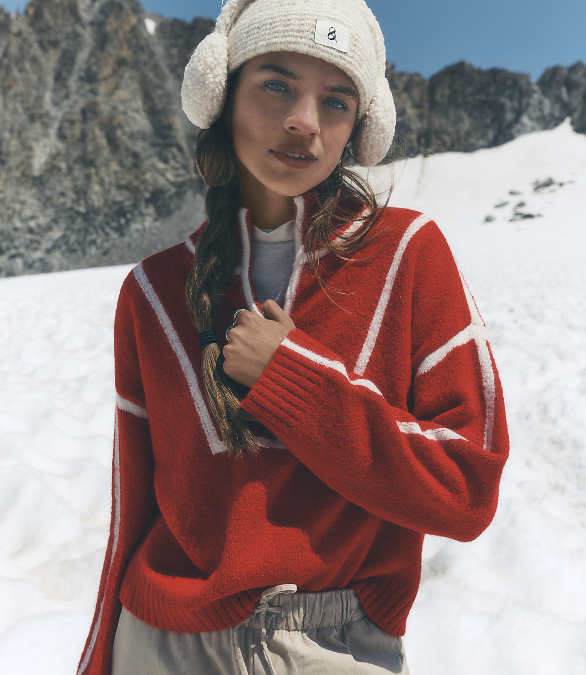 A woman wearing a red patterned sweater and a white beanie stands against a backdrop of snowy slopes and rugged mountains.
