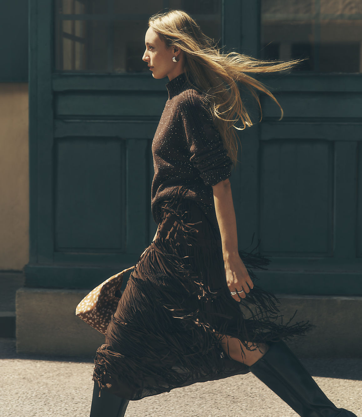 A blonde woman in a brown knit top and fringed skirt walks past a dark green paneled wall.