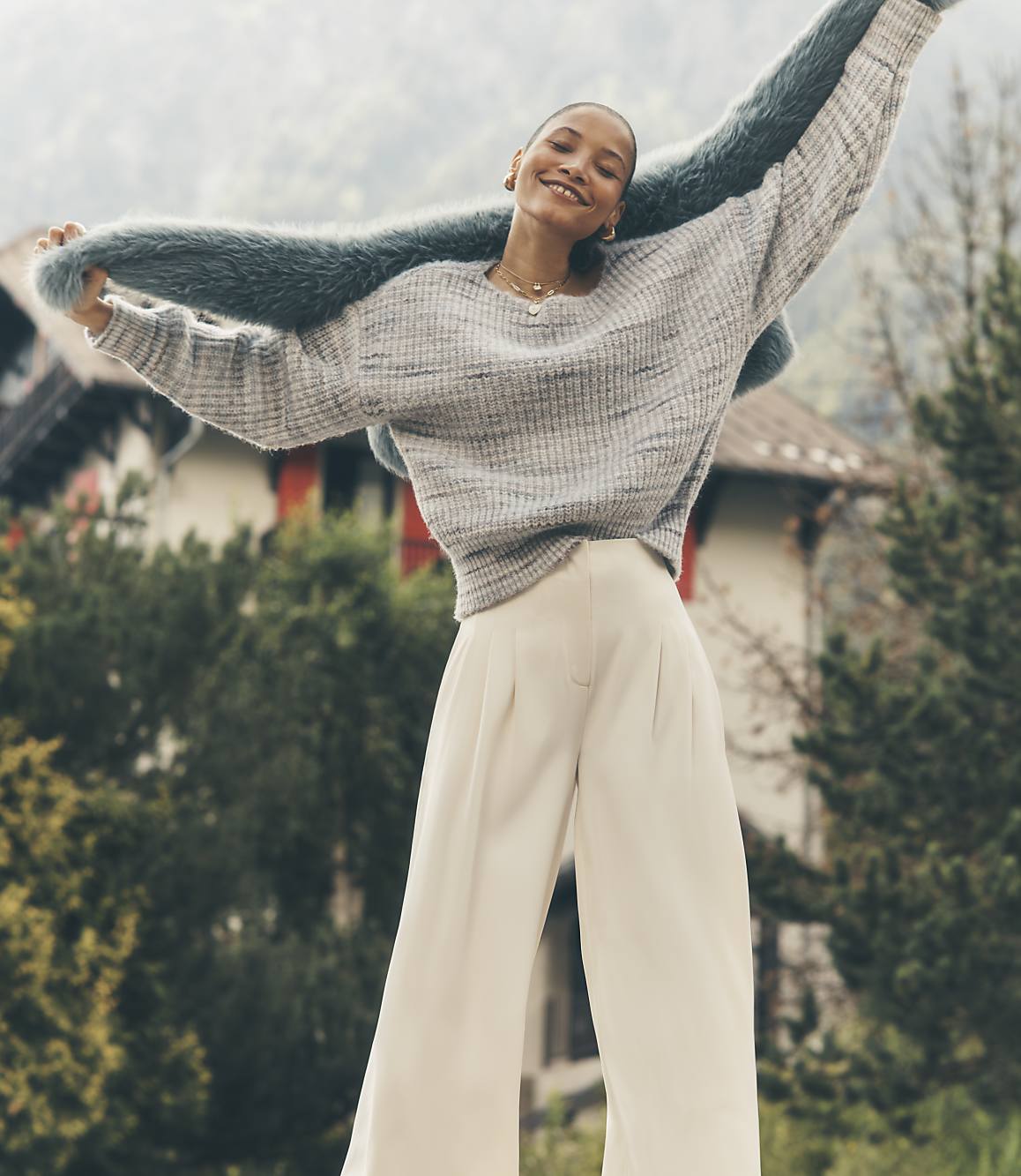 A smiling woman wearing a grey sweater and white trousers holds a blue faux-fur shawl aloft against a backdrop of a traditional house and misty mountains.