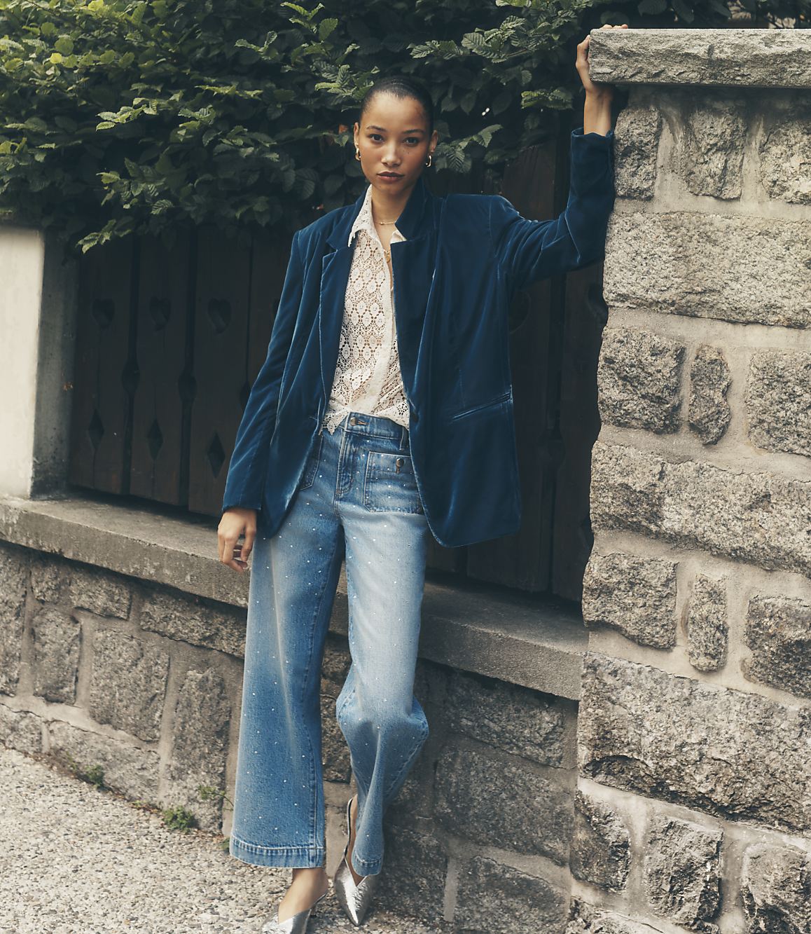 A woman wearing a blue velvet blazer and shimmering wide-leg jeans leans against a stone wall with lush green foliage in the background.