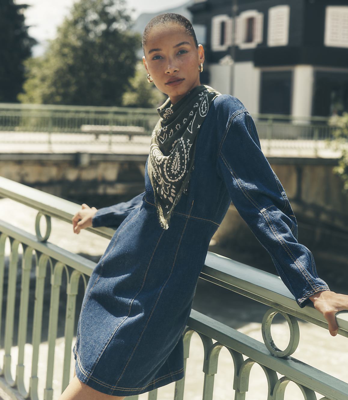 A woman in a denim dress and patterned scarf leans against a green railing with a blurred cityscape and greenery in the background.