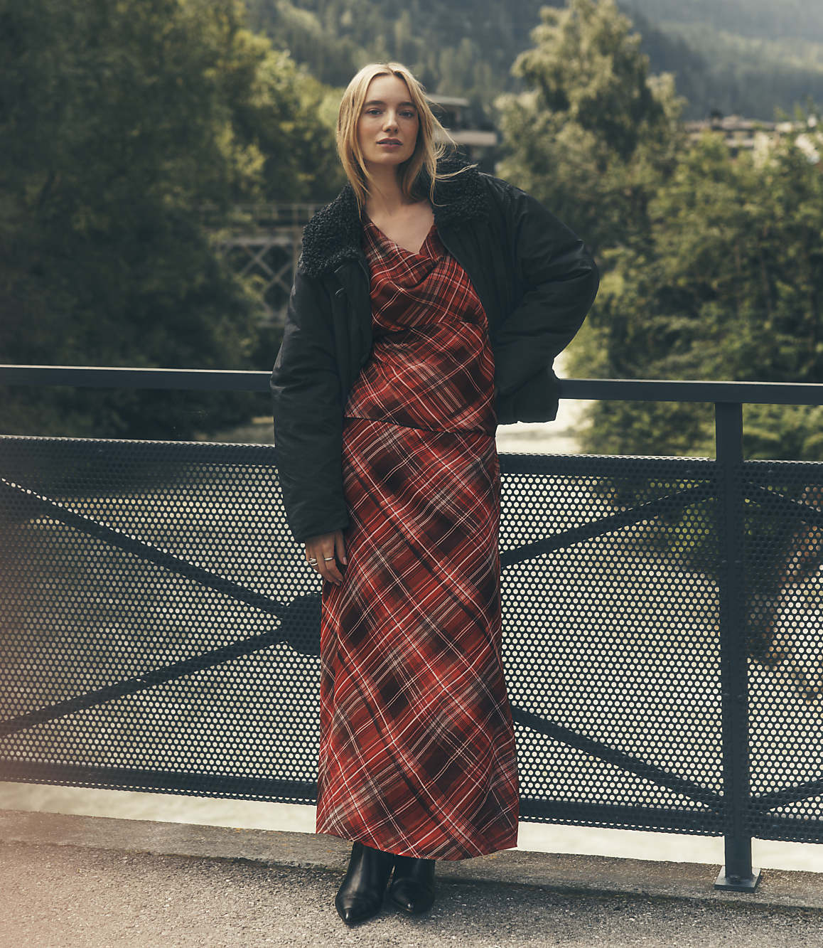 A woman wearing a red plaid dress and a black jacket stands against a metal railing with a lush, green forest in the background.