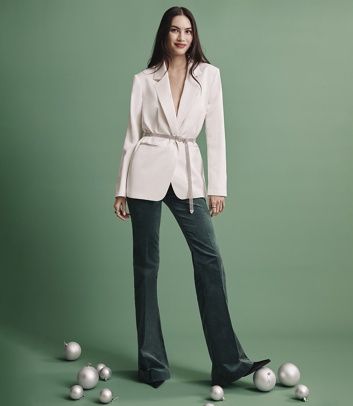 A woman wearing a white blazer and dark green trousers stands among silver ornaments against a solid green background.