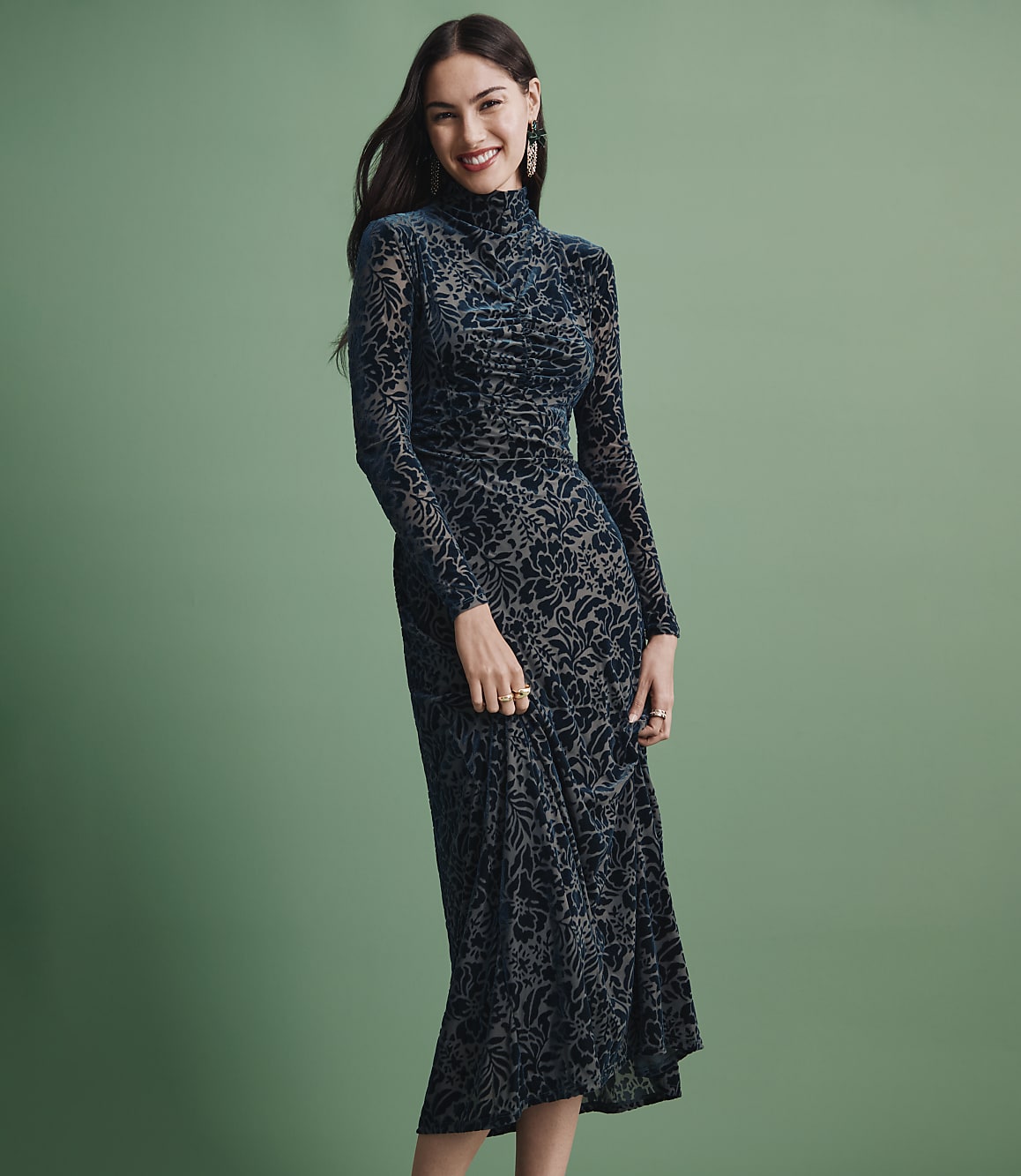 A smiling woman wearing a long-sleeved, dark teal patterned dress stands against a solid sage green background.