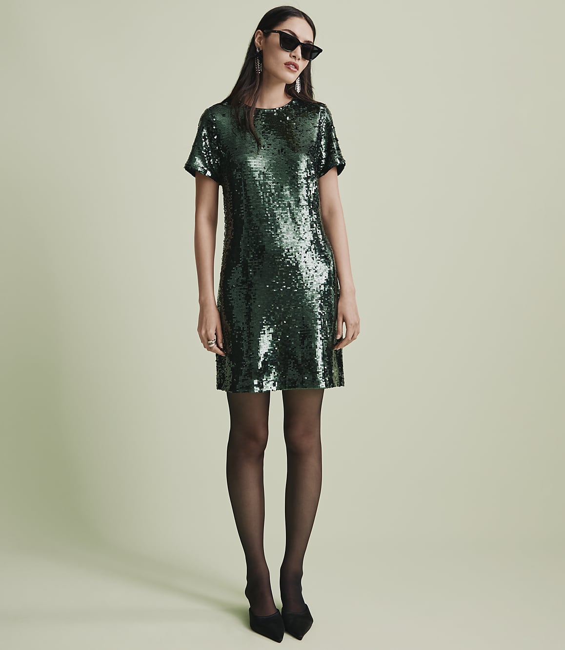 A woman wearing a green sequined dress, black tights, and sunglasses stands against a plain light green background.