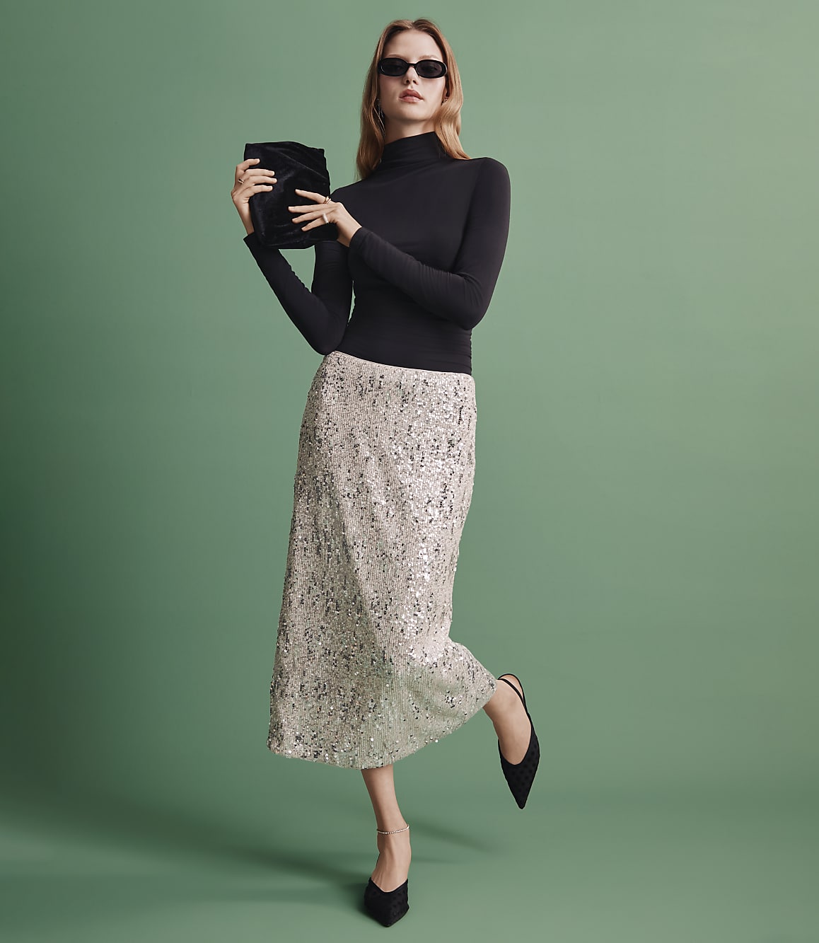 A woman wearing a black turtleneck, a shimmering silver sequined skirt, and sunglasses stands against a solid green background.