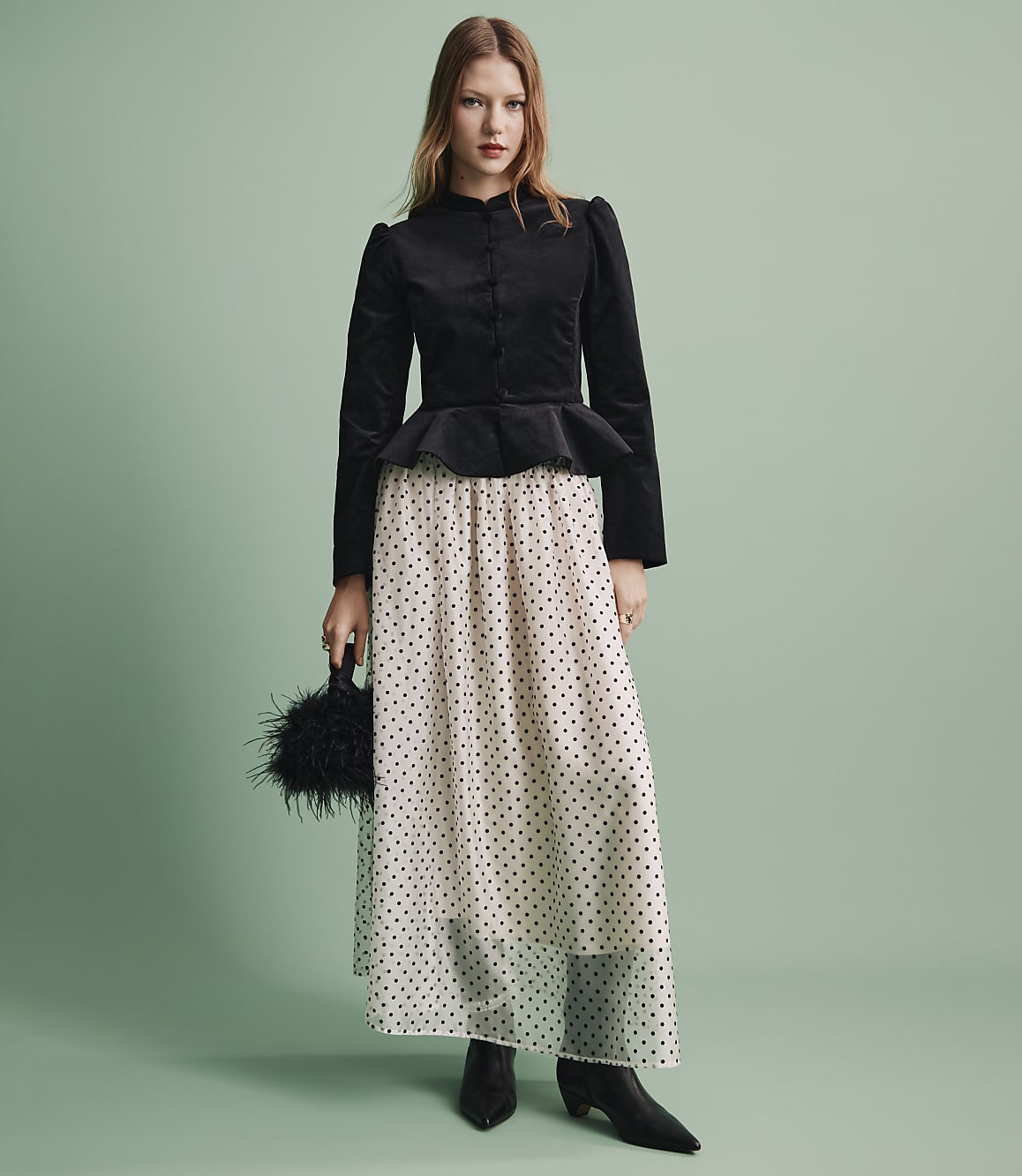 A woman wearing a black velvet peplum top and a sheer polka-dot skirt stands against a solid light green background.