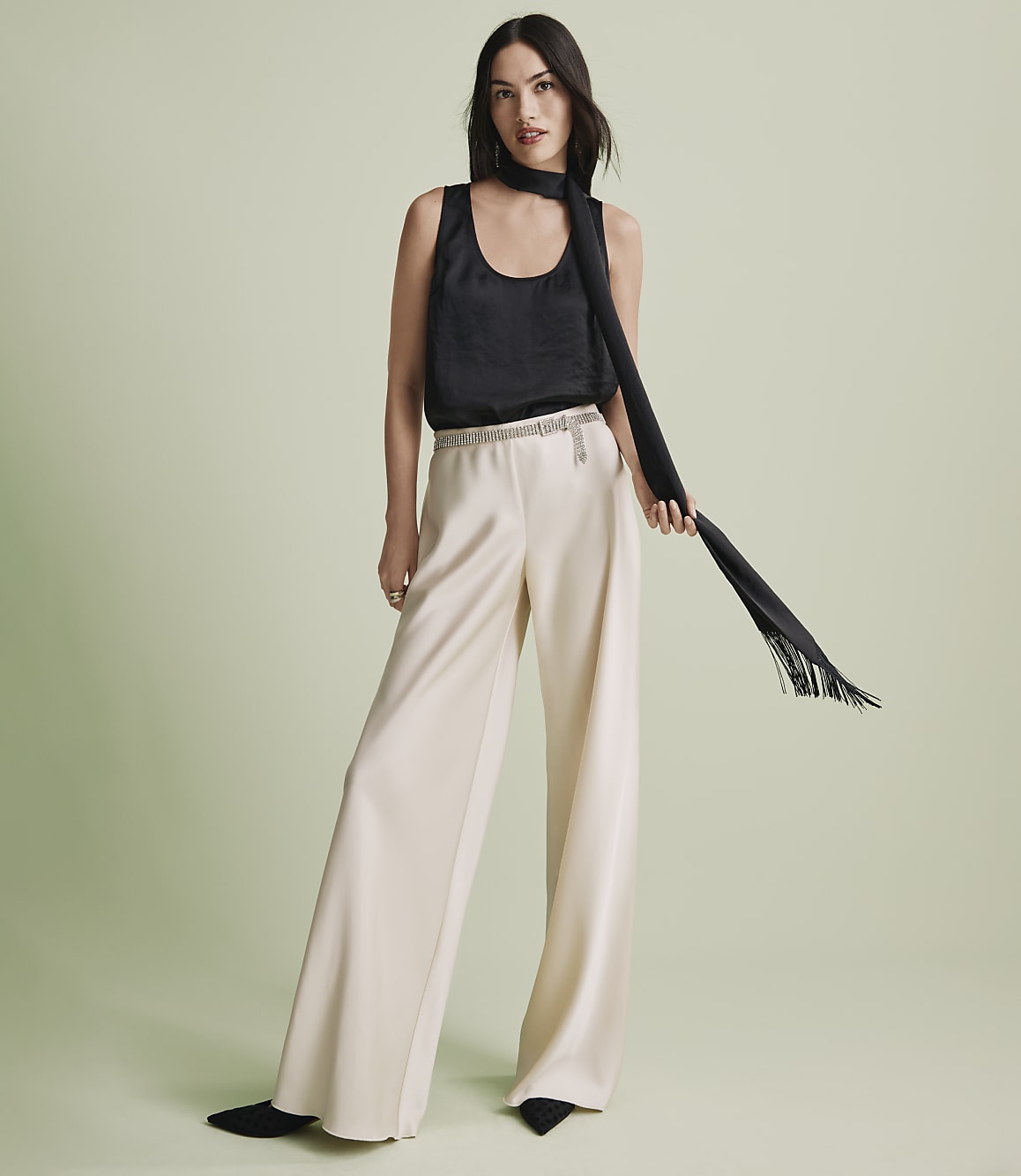 A woman wearing a black sleeveless top, wide-leg cream trousers, and a long black scarf stands against a plain light green background.