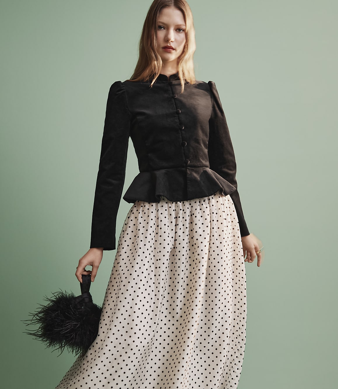 A woman wearing a black peplum top and a polka-dot skirt stands against a plain light green background.