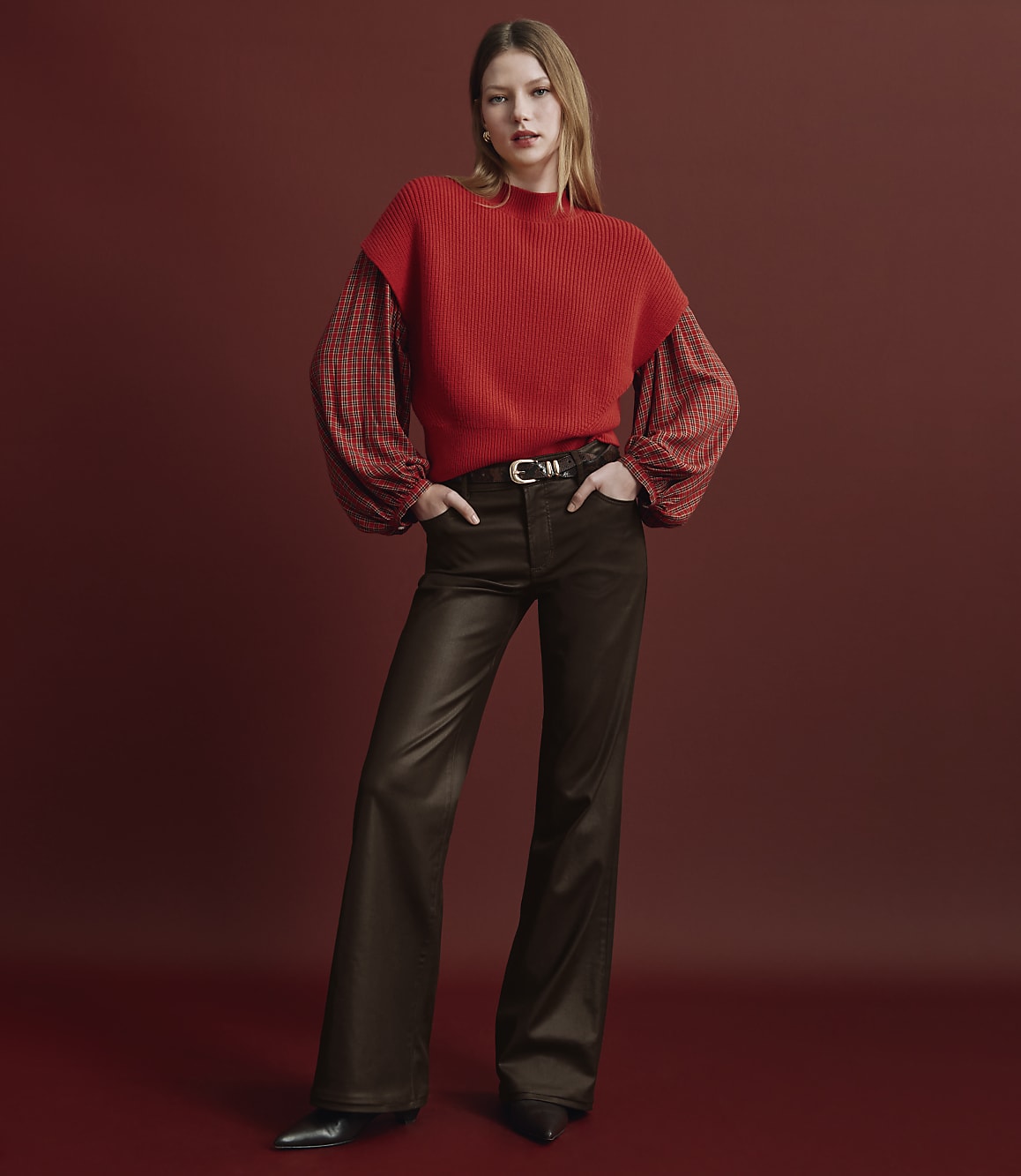 A woman wearing a red sweater and brown flared trousers stands against a solid red background.