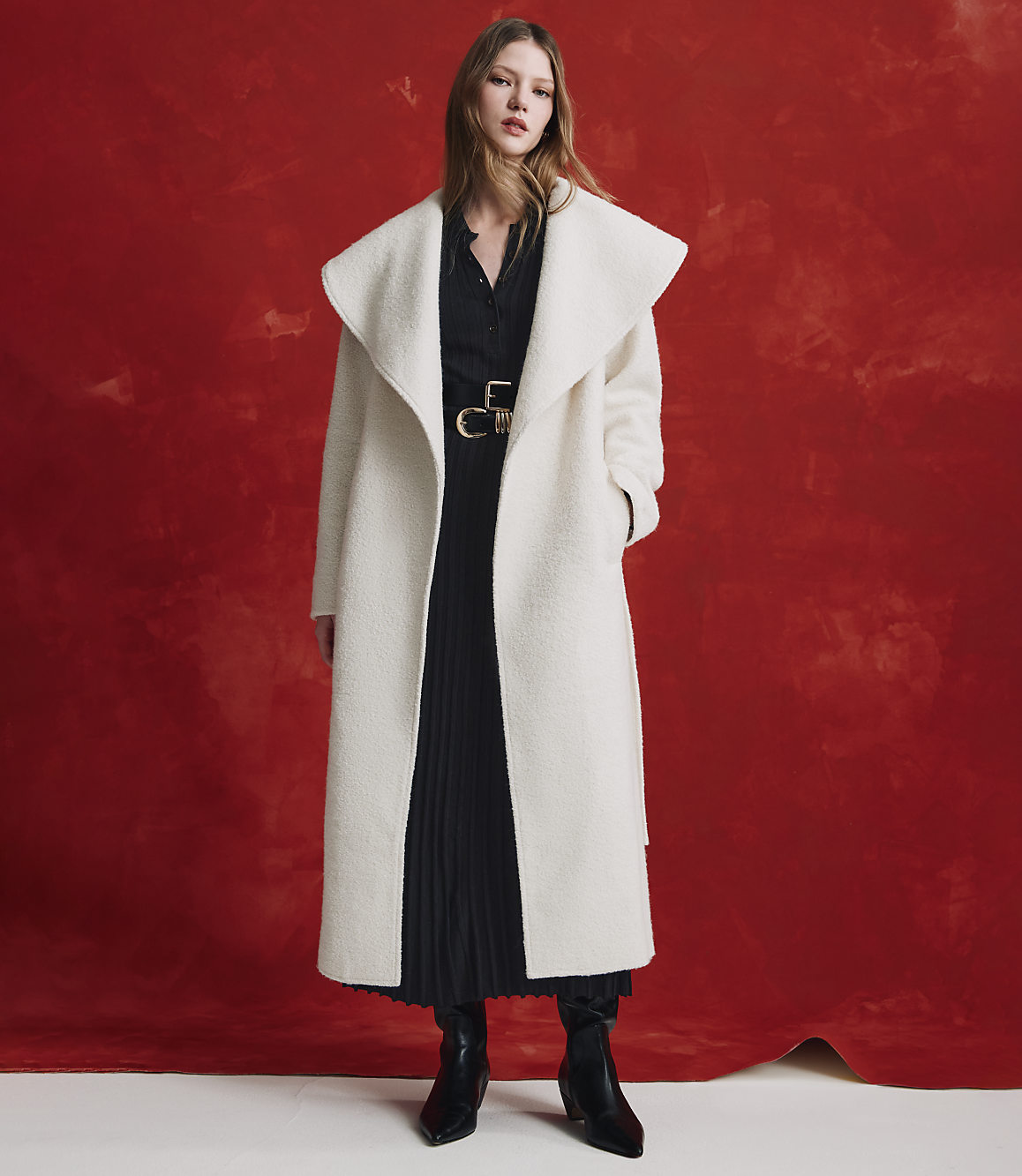 A woman wearing a long white coat over a black pleated dress and black boots stands against a textured red background.