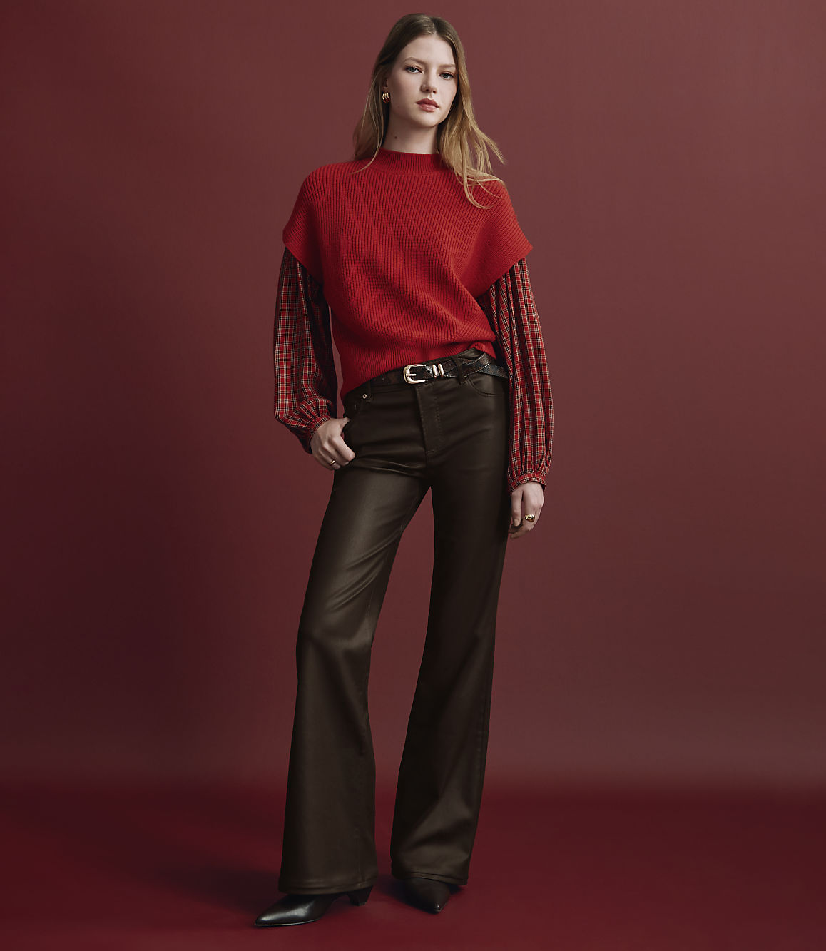 A woman wearing a red sweater and brown flared trousers stands against a solid red background.