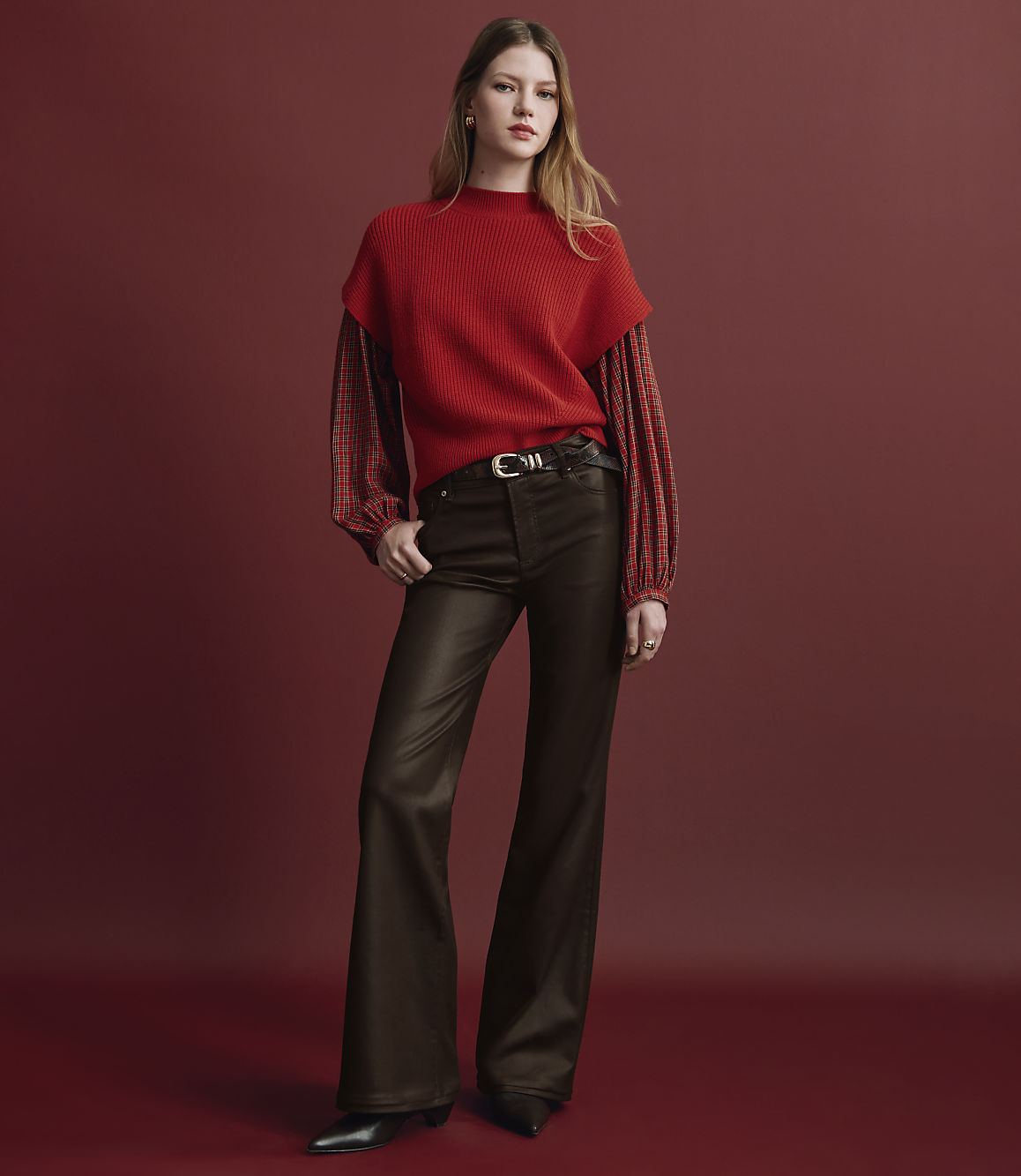 A woman wearing a red layered top and brown flared trousers stands against a solid red background.