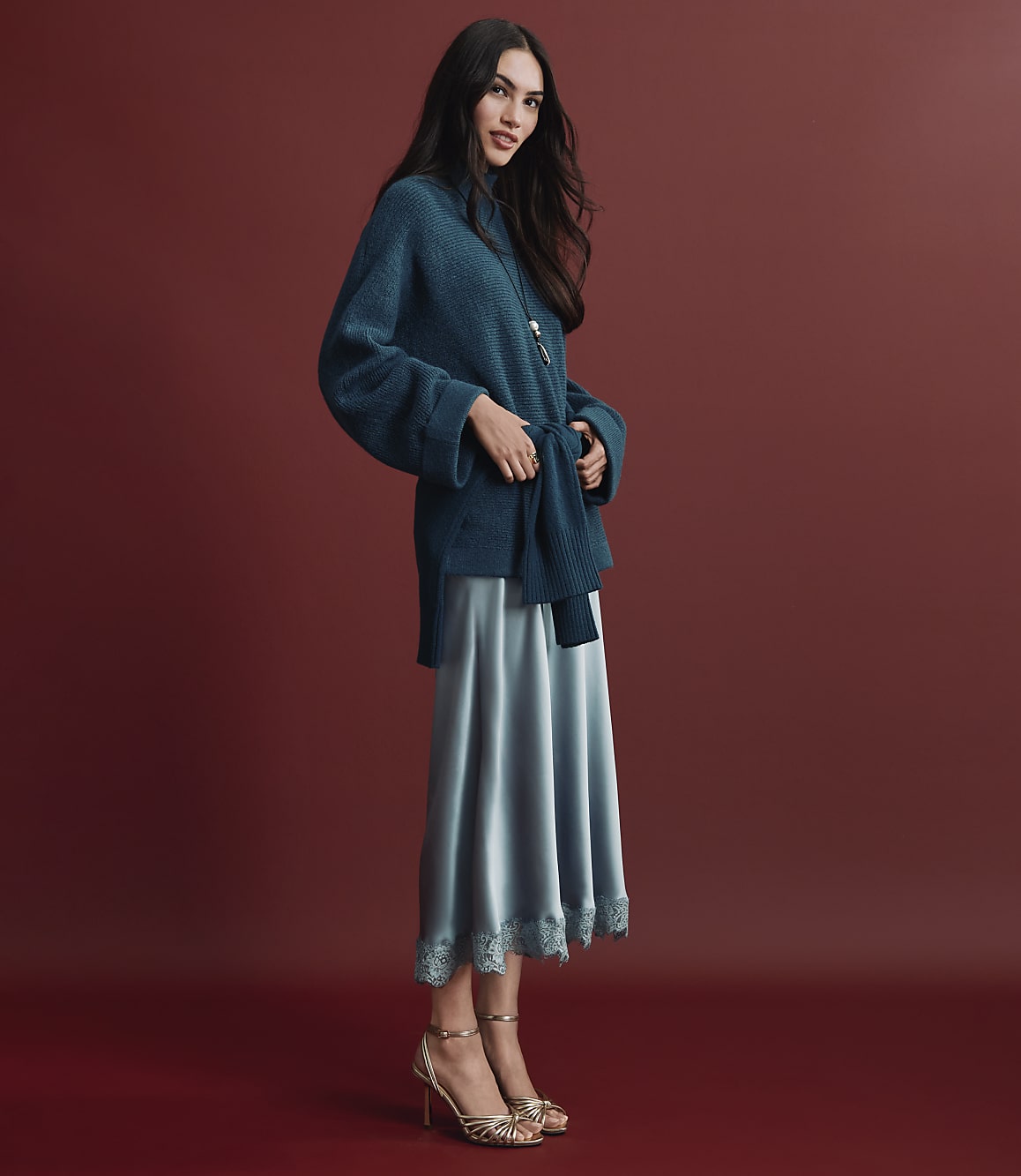 A woman wearing a teal knit sweater and a shimmering light blue satin skirt stands against a solid deep red background.