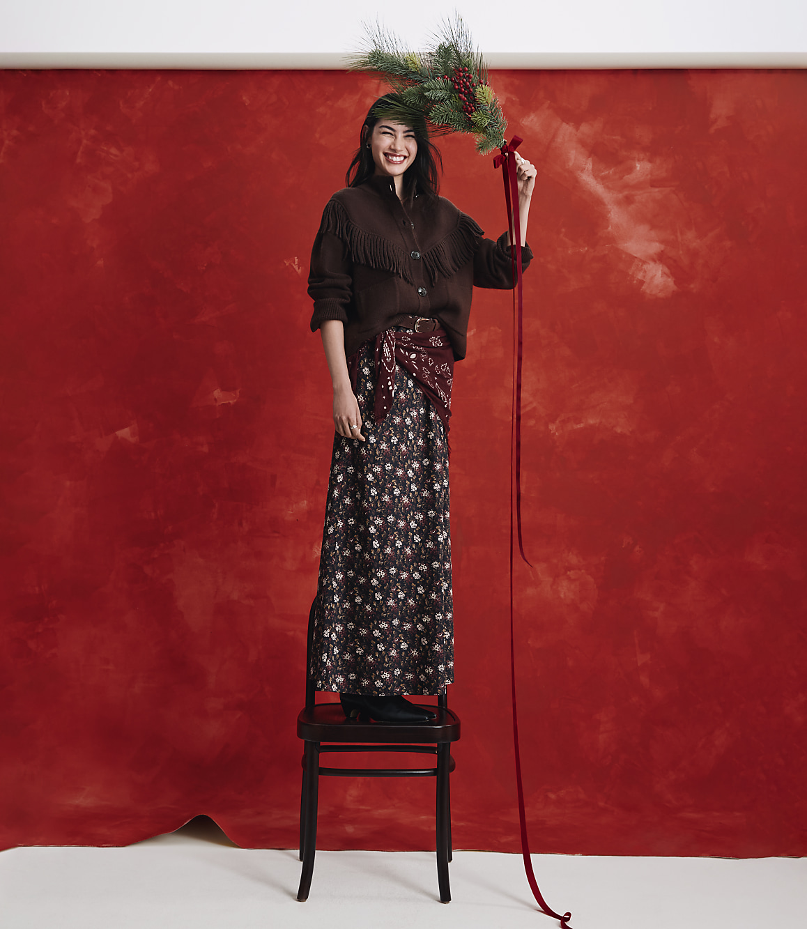 A smiling woman wearing a brown fringed cardigan and a floral skirt stands on a wooden chair against a textured red backdrop while holding a pine branch.