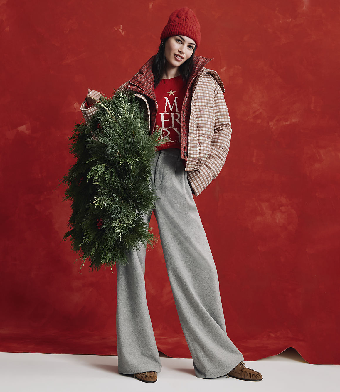 A woman wearing a red beanie, a checkered jacket, and wide-leg grey trousers holds a large evergreen wreath against a textured red background.