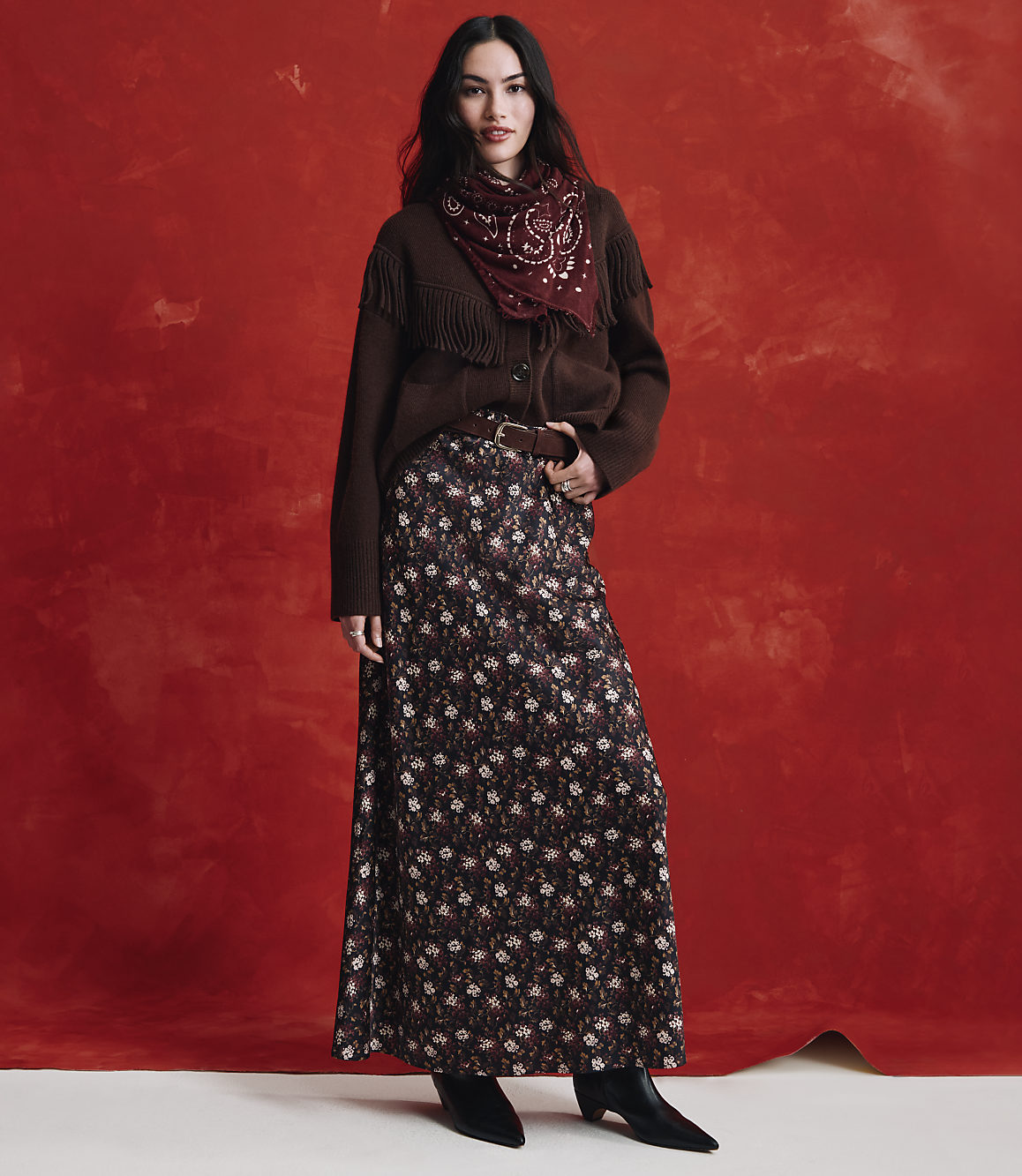 A woman wearing a brown cardigan, a red patterned scarf, and a long floral skirt stands against a textured red background.