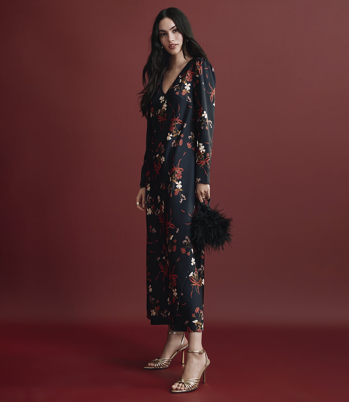 A woman wearing a long-sleeved floral black dress and gold heels stands against a solid red background.
