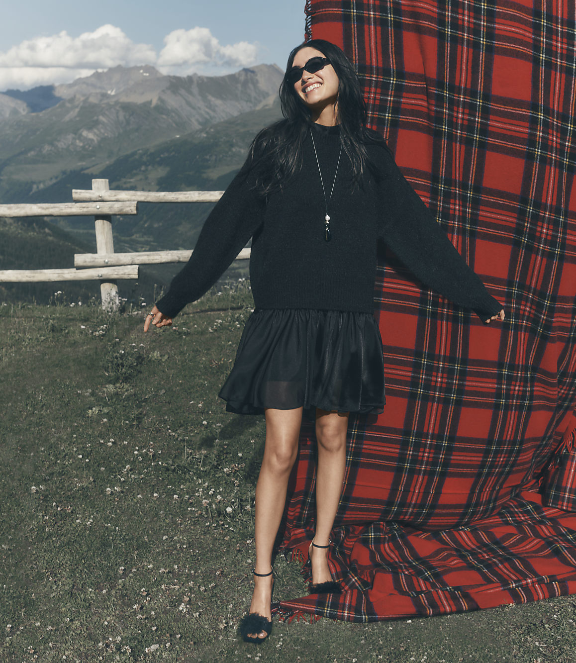 A woman in a black outfit stands before a large red plaid backdrop on a grassy hill with mountains in the distance.