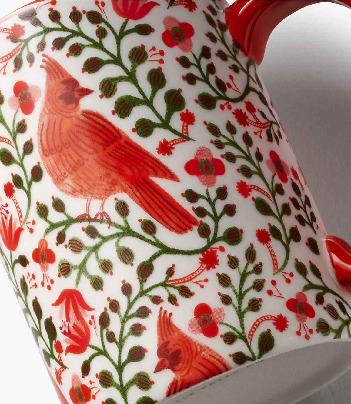 A red cardinal and floral patterns decorate the foreground of a white ceramic mug against a plain white background.
