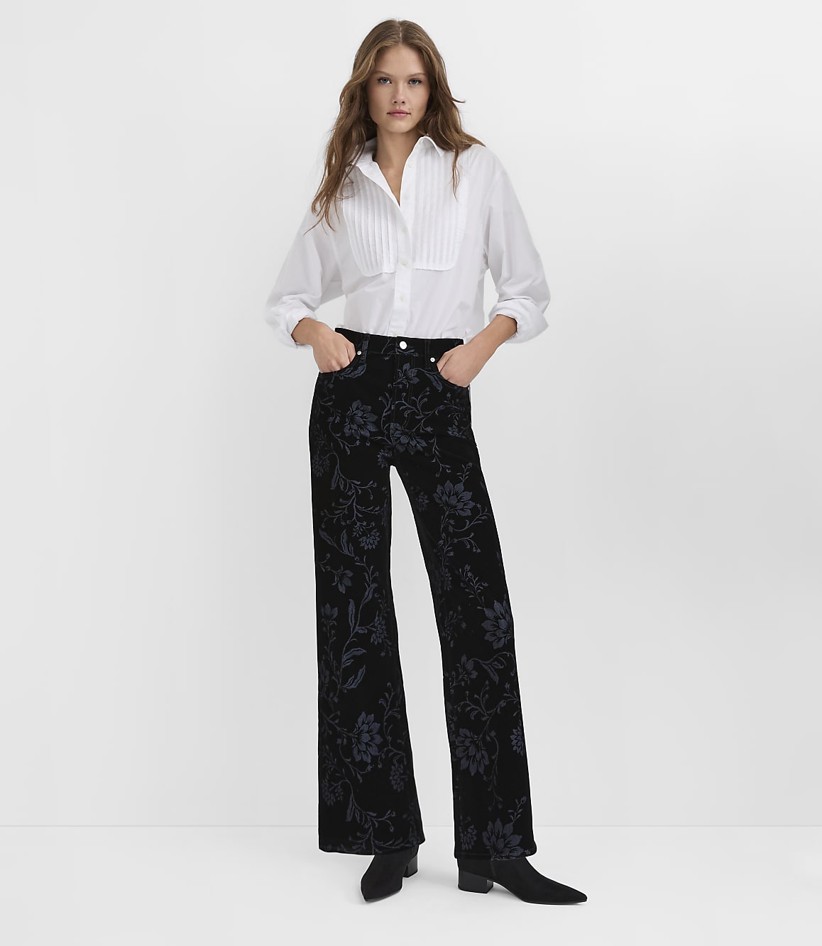 A woman wearing a white pleated blouse and floral black velvet trousers stands against a plain white background.