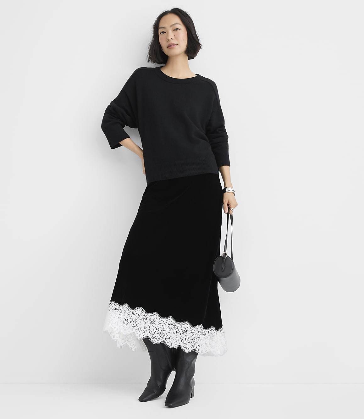 A woman wearing a black sweater, a long black skirt with white lace trim, and black boots stands against a plain white background.