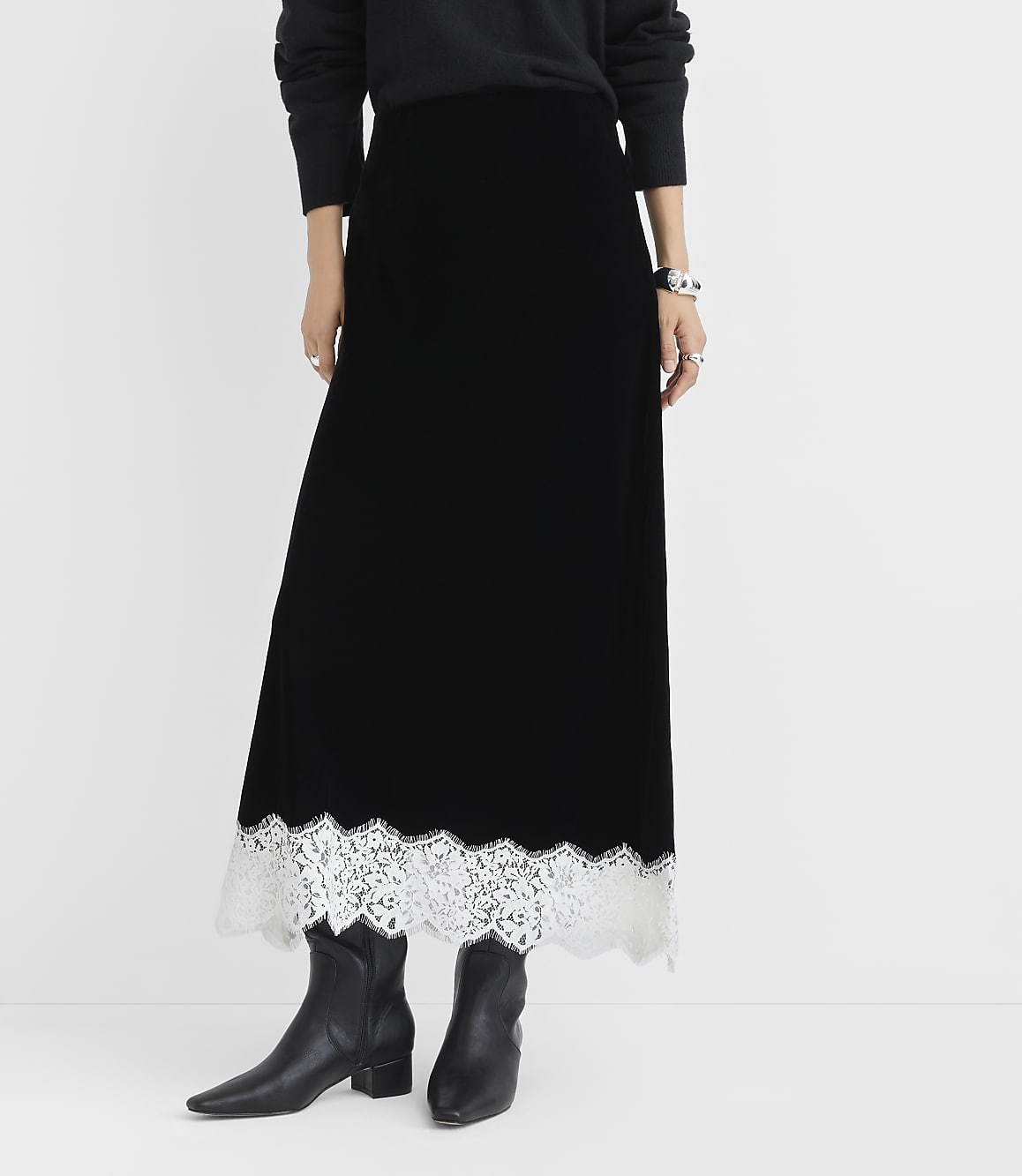 A person wearing a black velvet maxi skirt with a white lace hem and black boots stands against a plain white background.