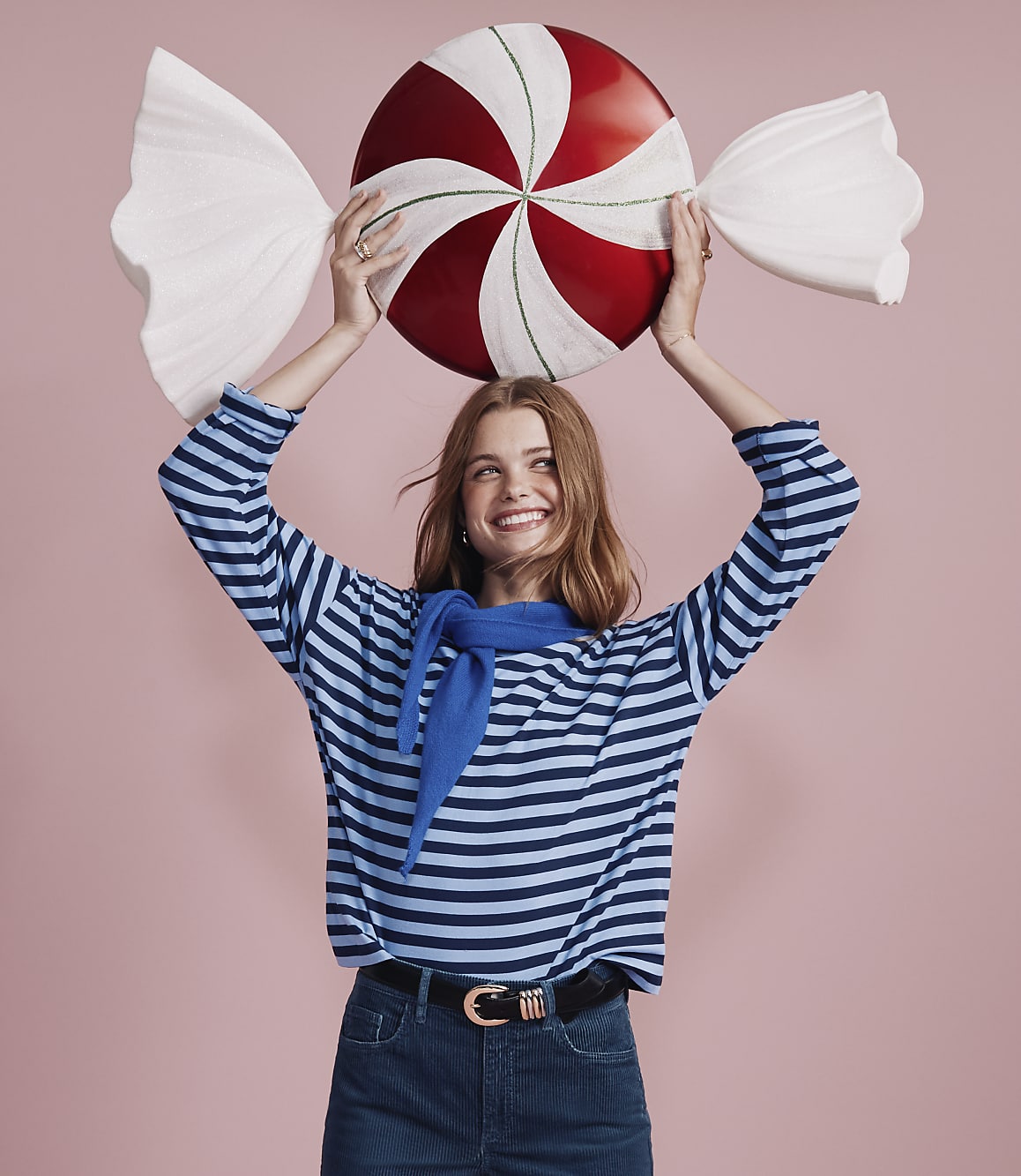 A smiling woman in a blue striped shirt holds a giant red and white peppermint candy against a solid pink background.