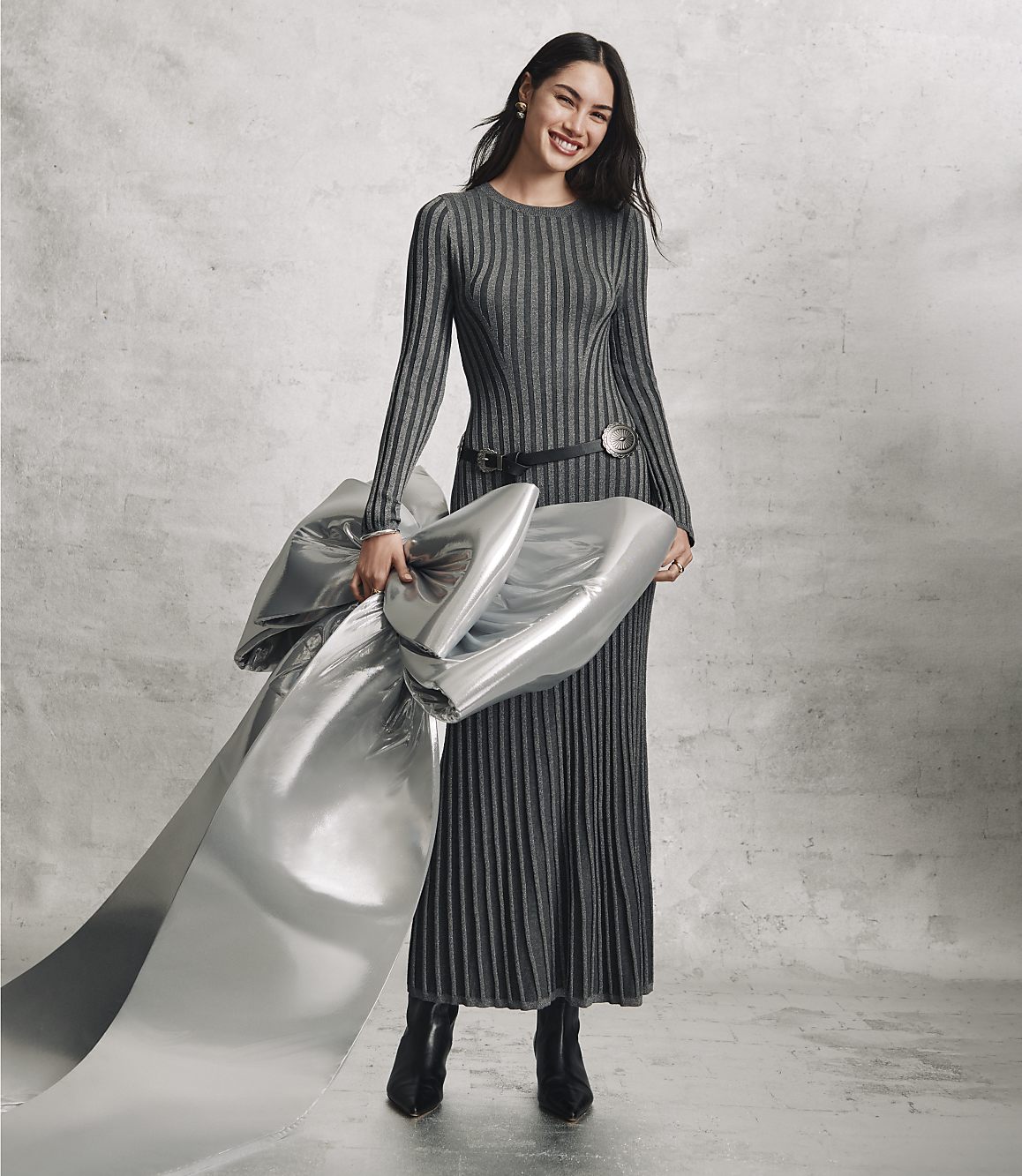 A smiling woman in a long, ribbed grey dress holds a large piece of shimmering silver fabric against a plain, textured grey concrete wall.