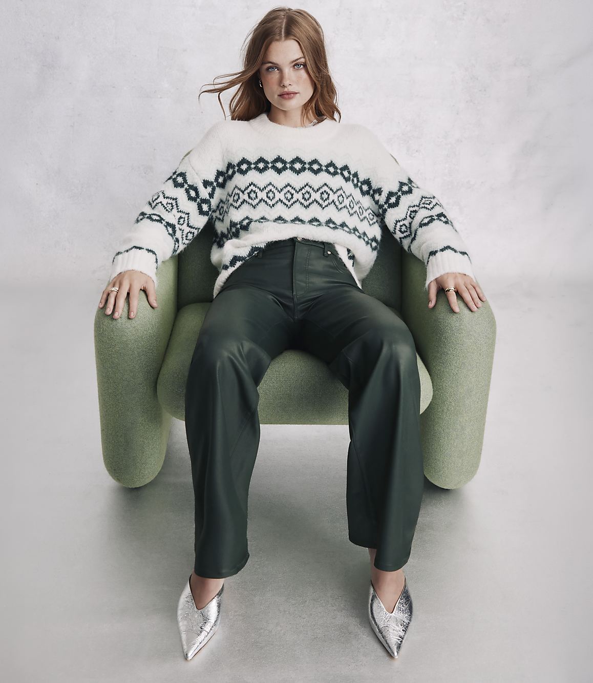 A woman wearing a patterned white sweater, dark green leather pants, and silver pointed shoes sits in a light green armchair against a plain grey background.
