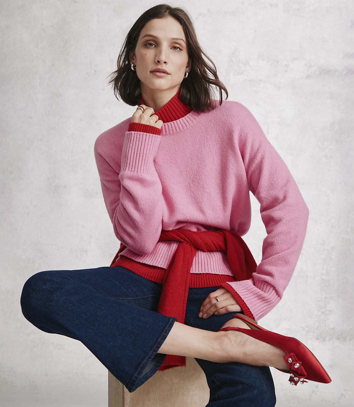 A woman wearing a pink sweater, red turtleneck, and blue jeans sits against a neutral, textured grey wall.
