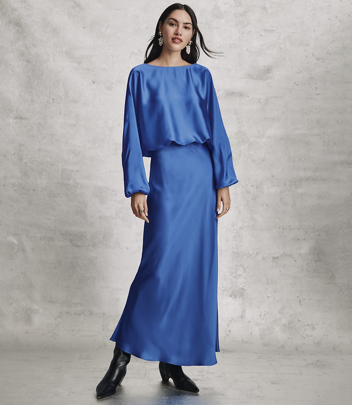 A woman wearing a long, royal blue satin dress and black boots stands against a textured grey concrete wall.