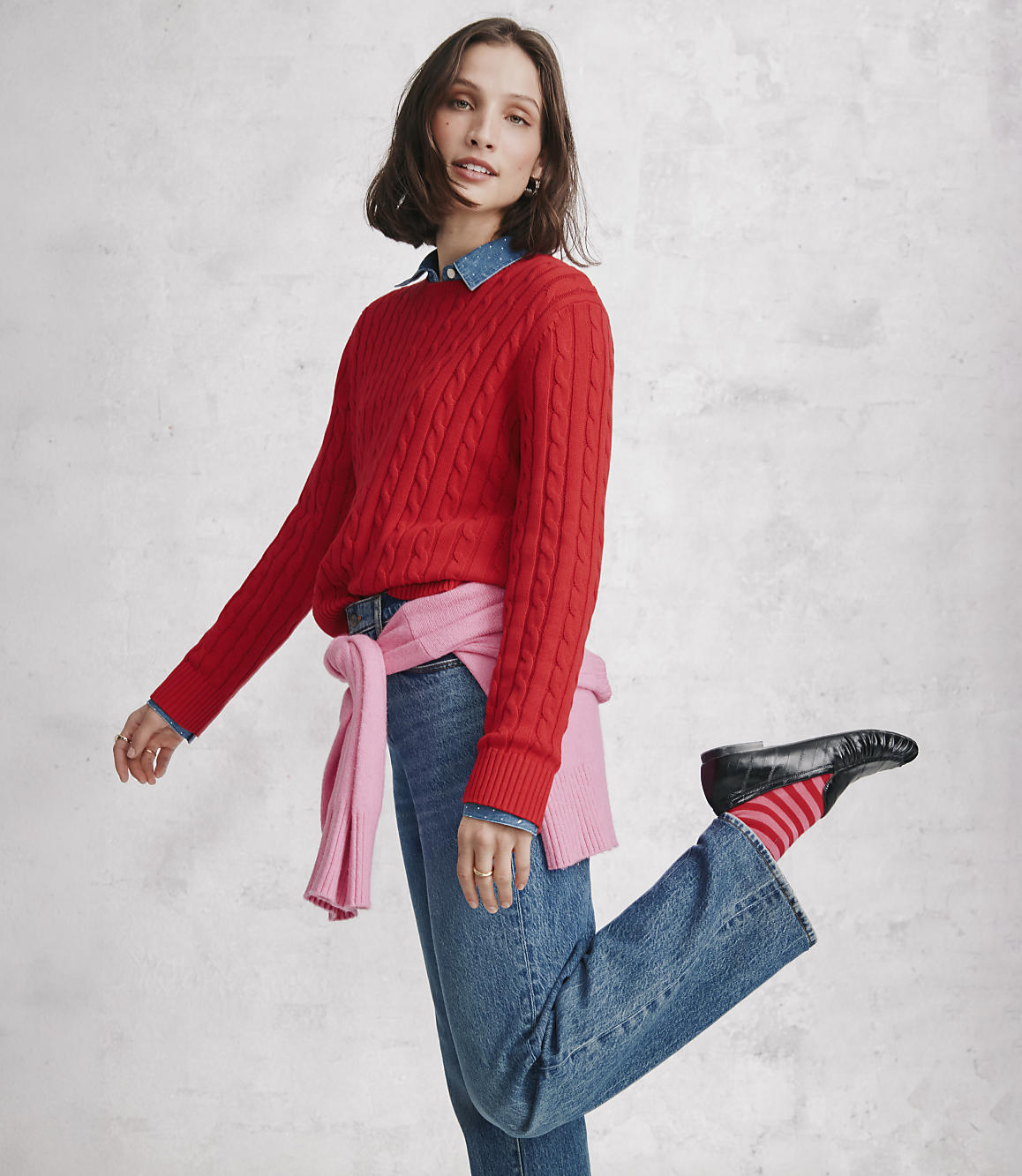 A woman wearing a red cable-knit sweater and blue jeans poses against a neutral, textured grey wall.