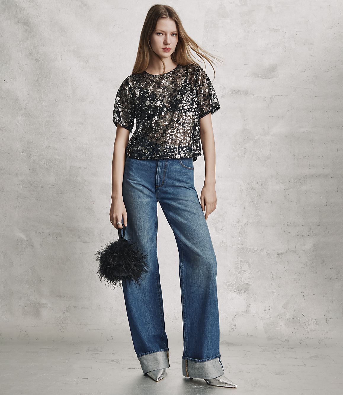 A woman wearing a sequined black top and wide-leg blue jeans stands against a textured grey wall.