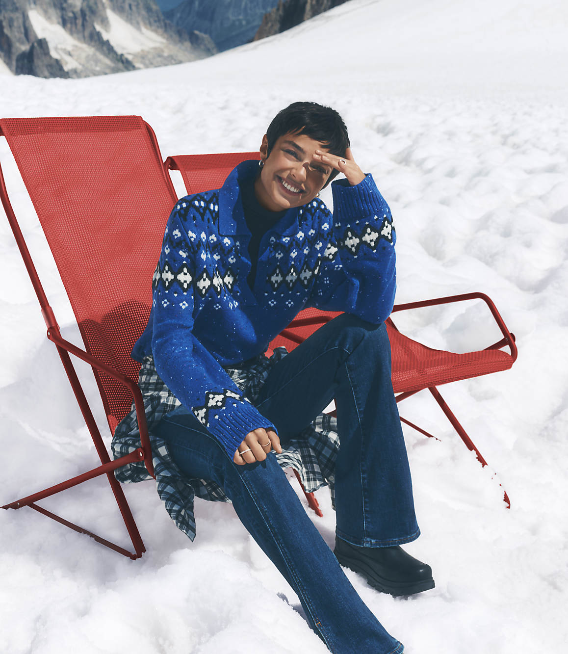 A smiling woman in a blue patterned sweater and jeans sits in a red lounge chair amidst a snowy landscape with mountains in the distance.