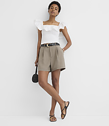 A woman in a white ruffled top and olive shorts stands against a plain white background, holding a small black purse. carousel Product Image 1